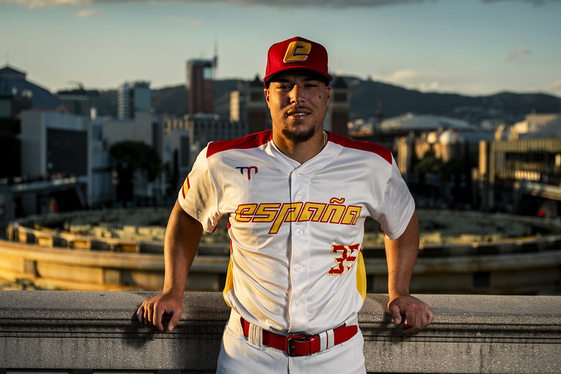 Spain catcher and captain Omar Hernández in the team’s white and red uniform, posed on a terrace with the Barcelona skyline behind him at sunset.