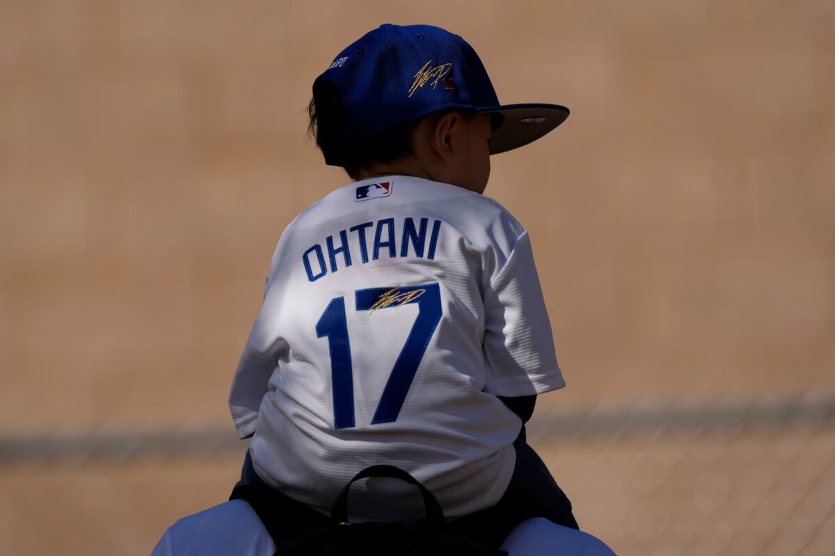 A child wearing a Los Angeles Dodgers' Shohei Ohtani jersey and cap rides on their father's shoulders on the first day of spring training baseball workouts at Camelback Ranch in Phoenix, Friday, Feb. 9, 2024(AP Photo/Carolyn Kaster)