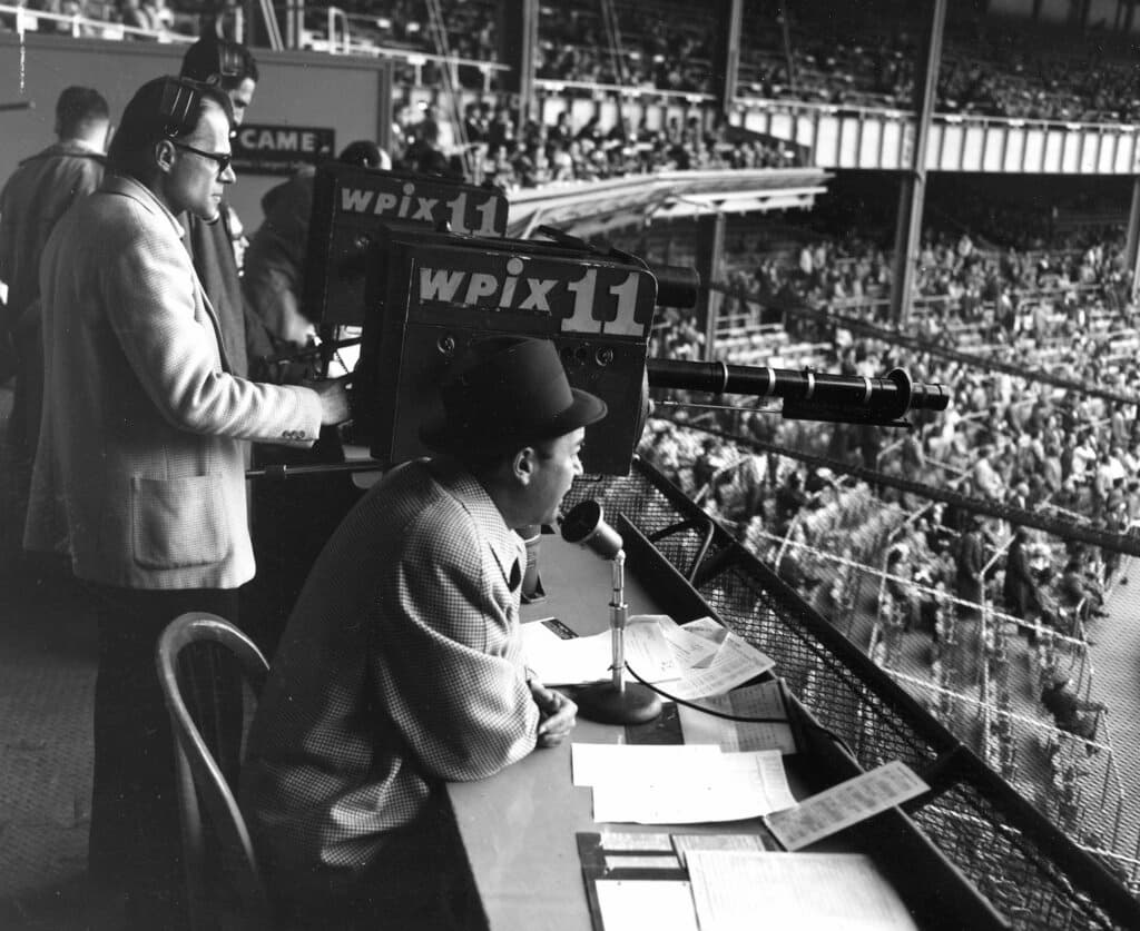 Title: Yankees Mel Allen Image ID: 560511014 Article: New York Yankees announcer Mel Allen, seated, reports the game between the Yankees and Baltimore Orioles from the television box behind home plate at Yankee Stadium on May 11, 1956. Behind the camera, center, is Jim Woods. (AP Photo/Robert Kradin)